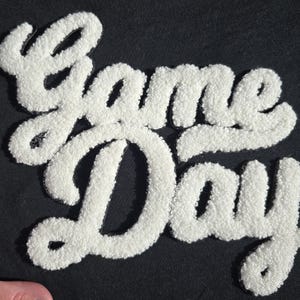 Peut inclure: Écusson blanc et texturé "Game Day" avec une police script, sur fond noir. Le patch a un aspect duveteux et en relief, créant un effet 3D. Les lettres sont liées, et le design est épuré et audacieux.