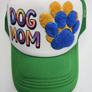 May include: A green and white trucker hat with the text "DOG MOM" in colorful tie-dye lettering. The hat features a paw print design in gold and blue, and a green brim.