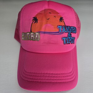 May include: A vibrant pink trucker hat with a curved peak and mesh back. The front displays a sunset graphic with palm trees and the words "Extra" and "Tanned & Tipsy" in glitter and blue text.