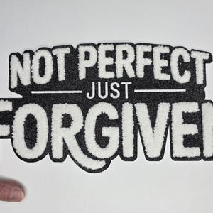May include: A black and white patch with the words "NOT PERFECT JUST FORGIVEN" in a bold, textured font. The patch has a black background with white lettering and a white border.
