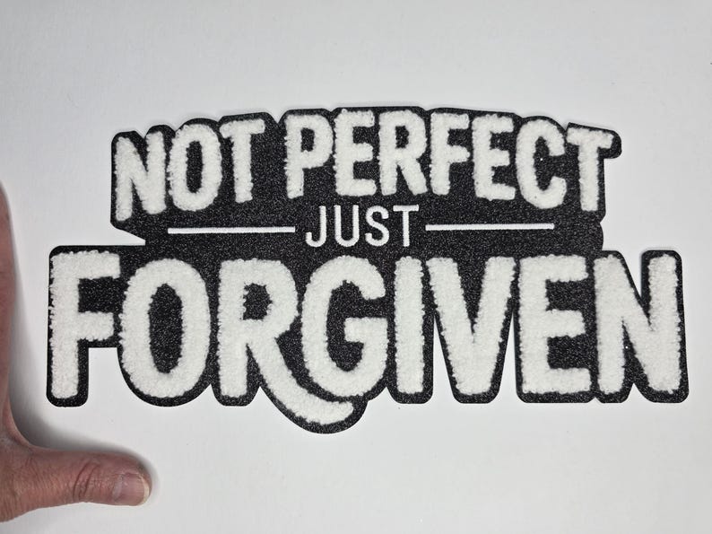 May include: A black and white patch with the words "NOT PERFECT JUST FORGIVEN" in a bold, textured font. The patch has a black background with white lettering and a white border.