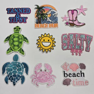 May include: A selection of embroidered patches with beach-themed designs. Includes patches with text that reads "TANNED & TIPSY", "BEACH BUM", and "SALTY". Other patches feature a turtle, crab, sun, and a pink cowboy boot.