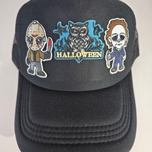 May include: Black trucker hat with a mesh back and a curved brim. The front features three embroidered patches: Jason Voorhees, an owl with the word "HALLOWEEN", and Michael Myers. The patches are in a cartoon style.