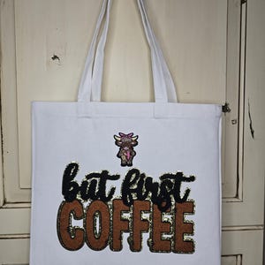 May include: White canvas tote bag with long handles. The bag features the phrase "but first COFFEE" in black and brown glitter lettering, with a small cartoon cow illustration above the text. The bag is hanging on a white hook.