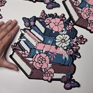 May include: Embroidered patches depicting a stack of books adorned with pink and white flowers and butterflies. The books are in shades of blue, pink, and brown, with white pages. The patches have a black border.