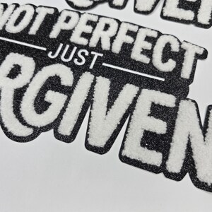 May include: Black and white embroidered patch with the words "NOT PERFECT JUST FORGIVEN". The text is in a bold, stylized font with a fuzzy, textured appearance. The patch has a black border.