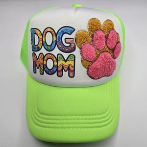May include: A neon green trucker hat with a white front panel. The front features the words "DOG MOM" in colorful bubble letters and a paw print design with pink and gold glitter accents. The hat has a curved brim.