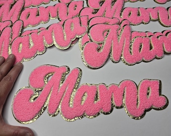 May include: Pink, fluffy "Mama" patches with gold glitter edging. The patches are in a cursive font and are scattered across a white surface. The patches are a bright pink colour.