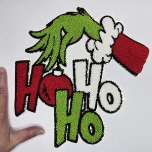 Peut inclure: Applique sur le thème de Noël avec une main verte et une main gantée rouge et blanche atteignant les mots "HO HO HO". Les lettres sont rouges, blanches et vertes avec une bordure dorée, et une boule rouge est entre les lettres "HO".