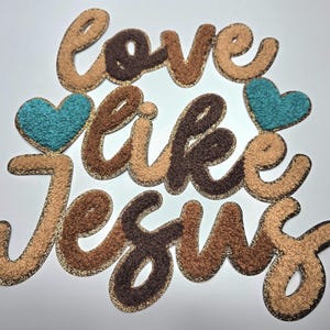 May include: A decorative felt applique with the words "love like Jesus" in various shades of brown, tan, and gold. Two teal blue hearts are placed on either side of the word "like". The letters have a textured, fuzzy appearance.