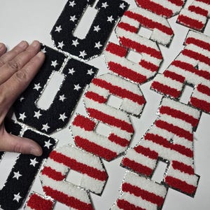 May include: Patches spelling "USA". The letter "U" is black with white stars, while the letters "S" and "A" have red and white stripes. The patches have a silver border.