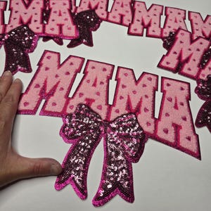 May include: Pink felt banner spelling "MAMA" with heart accents, and sequined bows. The letters are outlined in a darker pink glitter. The bows are a darker pink with sequins.