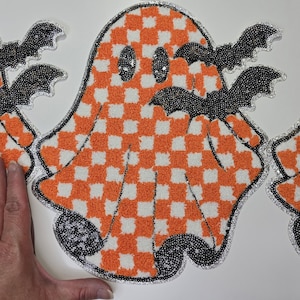May include: Halloween decoration in the shape of a ghost, with an orange and white chequered pattern. The ghost has sequined eyes and is decorated with black sequined bats. The edges are outlined with silver sequins.