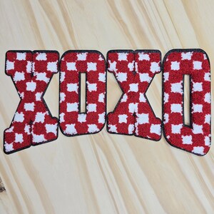 May include: Four embroidered letters spelling "XOXO" in a red and white chequered pattern. The letters have a black border and are set against a light wood grain background.