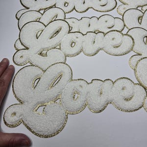 May include: White, fluffy fabric patches spelling "Love" in a cursive font. The edges and outlines of the letters are trimmed with gold glitter. The patches are arranged on a white surface.