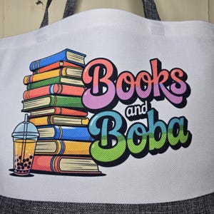 May include: White tote bag with a colorful graphic of a stack of books, a boba drink, and the words "Books and Boba" in a retro font. The bag has gray handles and a gray bottom.