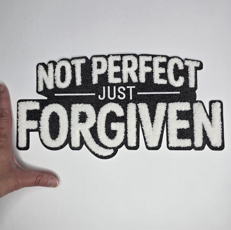 May include: A black and white sign with the words "NOT PERFECT" above "JUST" and "FORGIVEN" in a bold, stylized font. The sign has a textured appearance, with the white letters standing out against the black background.