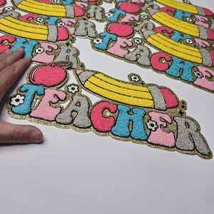Peut inclure: Écussons brodés avec le mot « TEACHER » dans une police rétro, avec un crayon jaune, une pomme rose et des motifs floraux. Les écussons ont une bordure dorée scintillante et sont de différentes couleurs, dont le rose, le bleu et le gris.
