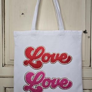 May include: White tote bag with two "Love" text designs in red and pink glitter. The bag has two white handles and is hanging against a white background.