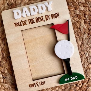 May include: A wooden picture frame with the word "DADDY" in white letters at the top. The phrase "You're the best, by par!" is engraved below. A golf ball and flag are on the right, with "#1 Dad" at the bottom.