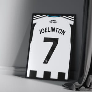 May include: Framed soccer jersey featuring the name "JOELINTON" and the number "7" in black on a white background. The jersey has black and white striped accents and the text "HOWAY THE LADS" at the top. The frame is black.