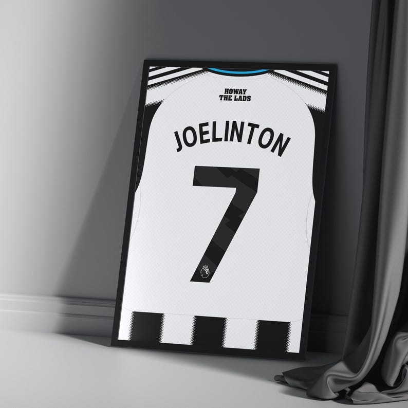 May include: Framed soccer jersey featuring the name "JOELINTON" and the number "7" in black on a white background. The jersey has black and white striped accents and the text "HOWAY THE LADS" at the top. The frame is black.