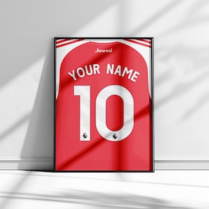 May include: Framed red and white Arsenal jersey print. The jersey features the number 10 in white, with the text "YOUR NAME" above. The Arsenal logo is visible, and the frame is black.
