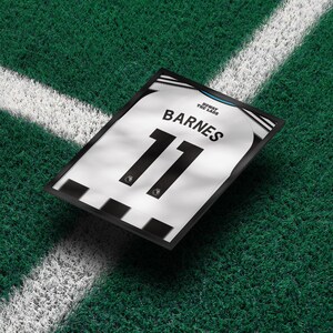May include: Framed soccer jersey with the name "BARNES" and the number "11" in black on a white and black striped jersey. The frame is black, and the background is green.