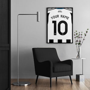 May include: A framed soccer jersey with the text "YOUR NAME" and the number "10" in black on a white background. The jersey is displayed on a wall above a black armchair. A silver floor lamp and a small side table with a vase of flowers are also in the room.