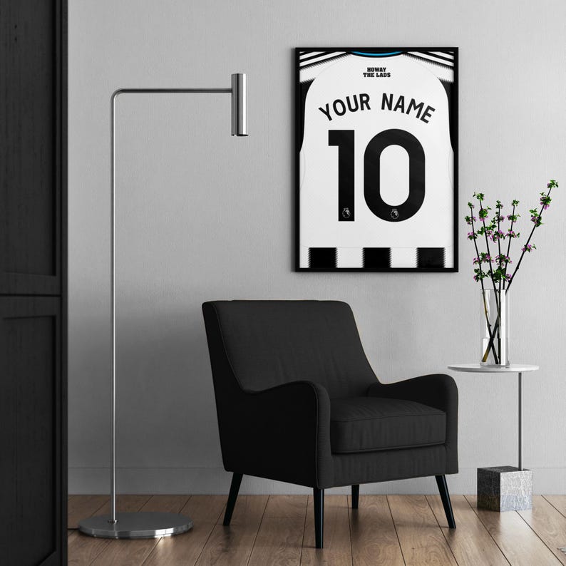 May include: A framed soccer jersey with the text "YOUR NAME" and the number "10" in black on a white background. The jersey is displayed on a wall above a black armchair. A silver floor lamp and a small side table with a vase of flowers are also in the room.