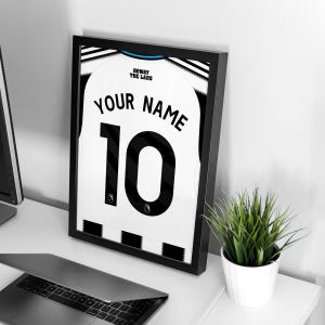 May include: Framed black and white soccer jersey print with the text "YOUR NAME" and the number "10". The jersey has a black and white striped design at the bottom and the words "HOWAY THE LADS" at the top.