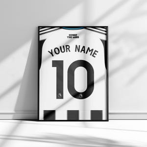 May include: Framed soccer jersey art print. The white jersey features a black number 10 and the text "YOUR NAME" above it. The words "HOWAY THE LADS" are at the top. The jersey is framed in a black frame.