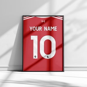 May include: Framed red jersey art with the text "YOUR NAME" above the number "10" in white. The jersey has white stripes and logos. The artwork is in a black frame and is displayed against a white wall.