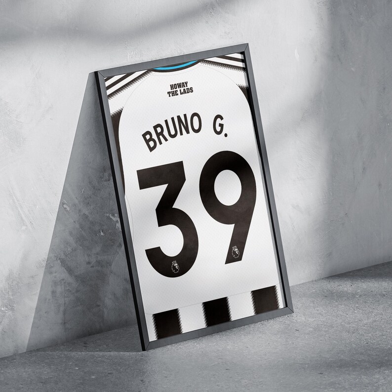 May include: Framed white and black soccer jersey with the name "Bruno G." and the number "39" in large black font. The jersey has a black frame and is leaning against a gray wall.