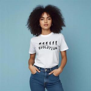 May include: White t-shirt with a black graphic depicting the evolution of a golfer. The word "EVOLUTION" is printed below the graphic in a curved font. The model is wearing blue jeans.