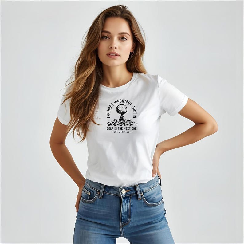 May include: White t-shirt with a black graphic of a golf ball on a tee, with the text "THE MOST IMPORTANT SHOT IN GOLF IS THE NEXT ONE - LET'S PAR TEE." The shirt is paired with blue jeans.
