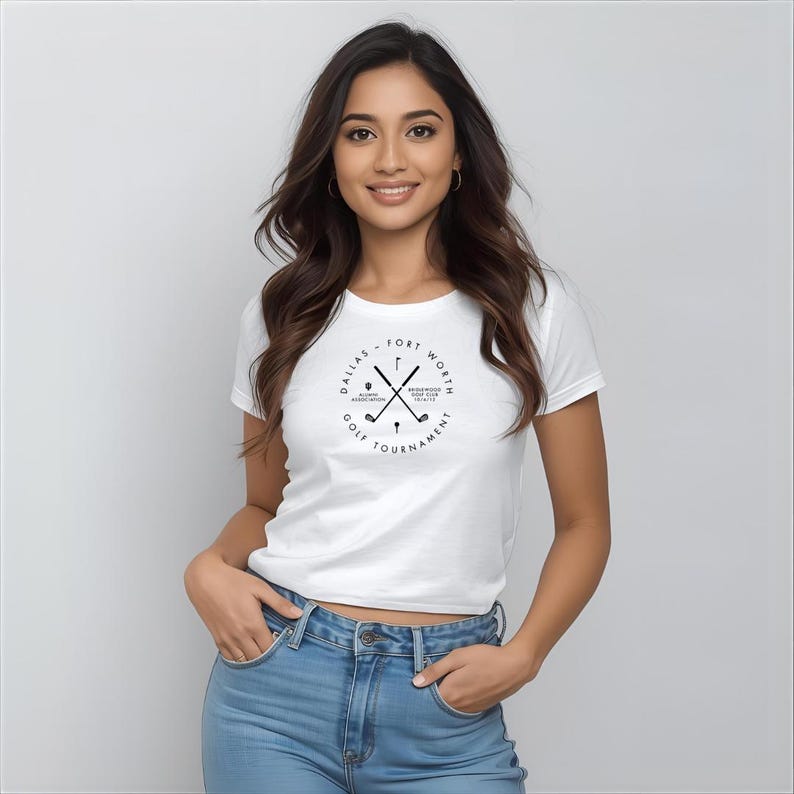 May include: White t-shirt with a black graphic featuring crossed golf clubs and text that reads "Dallas - Fort Worth Golf Tournament". The shirt is a crew neck style and is being worn.