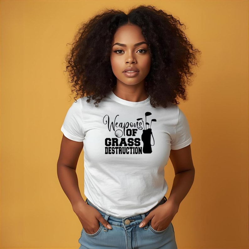 May include: White t-shirt with the text "Weapons of Grass Destruction" in black. The graphic includes a golf ball, golf clubs, and a golf bag. The model is wearing blue jeans.