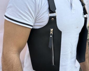 May include: A black leather shoulder holster bag featuring two vertical pockets with silver-toned metal zippers and adjustable straps with metal buckles. The design includes stitched edges and a structured shape that rests against the chest area.