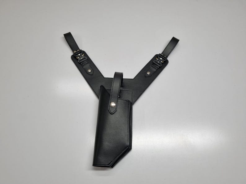 May include: A black leather shoulder holster featuring a Y-shaped harness with adjustable buckles and metal rivets. The holster includes a vertical pouch with a snap-button strap closure, designed to secure an item firmly in place against the body.