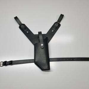 May include: A black leather shoulder holster system featuring a vertical gun holster attached to a Y-shaped harness with adjustable straps, metal buckles, and a horizontal waist belt with a silver-toned metal buckle and multiple adjustment holes.