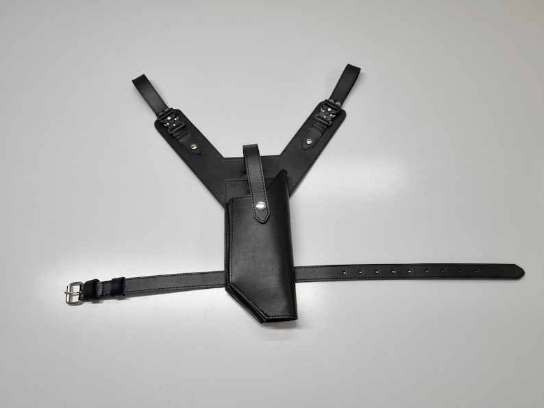 May include: A black leather shoulder holster system featuring a vertical gun holster attached to a Y-shaped harness with adjustable straps, metal buckles, and a horizontal waist belt with a silver-toned metal buckle and multiple adjustment holes.