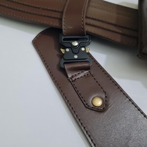 Puede incluir: Primer plano de un cintur&oacute;n de cuero marr&oacute;n con una hebilla de metal negro y un remache dorado. El cintur&oacute;n est&aacute; hecho de cuero liso marr&oacute;n oscuro y presenta costuras detalladas.