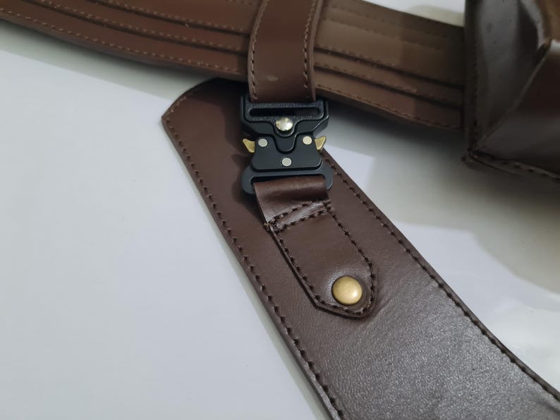 Puede incluir: Primer plano de un cintur&oacute;n de cuero marr&oacute;n con una hebilla de metal negro y un remache dorado. El cintur&oacute;n est&aacute; hecho de cuero liso marr&oacute;n oscuro y presenta costuras detalladas.