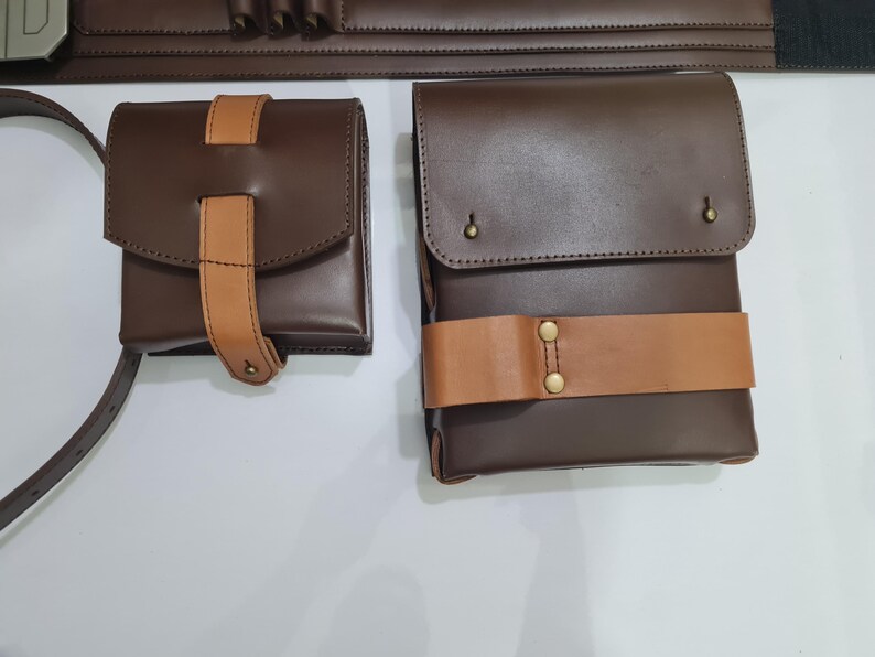 Puede incluir: Dos bolsas de cuero marr&oacute;n con correas de cuero color canela y herrajes de lat&oacute;n. Una bolsa es cuadrada con una solapa y una correa vertical. La otra es rectangular con una correa horizontal. Se ve un cintur&oacute;n a juego.