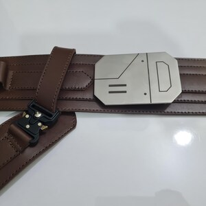 Puede incluir: Un cintur&oacute;n de cuero marr&oacute;n con una hebilla plateada con un dise&ntilde;o geom&eacute;trico. El cintur&oacute;n tiene una serie de peque&ntilde;os lazos y un cierre negro. El cintur&oacute;n est&aacute; dise&ntilde;ado con una serie de l&iacute;neas horizontales.