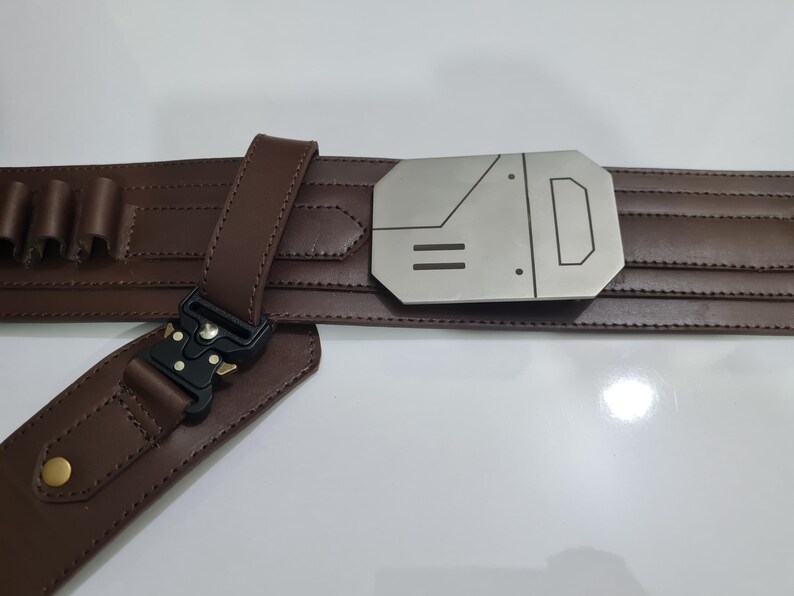 Puede incluir: Un cintur&oacute;n de cuero marr&oacute;n con una hebilla plateada con un dise&ntilde;o geom&eacute;trico. El cintur&oacute;n tiene una serie de peque&ntilde;os lazos y un cierre negro. El cintur&oacute;n est&aacute; dise&ntilde;ado con una serie de l&iacute;neas horizontales.