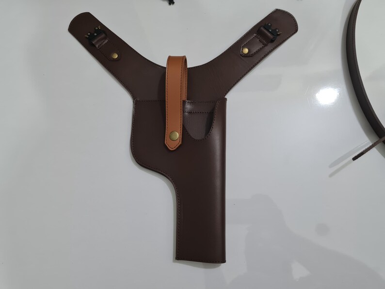 Puede incluir: Una funda de hombro de cuero marr&oacute;n con una funda vertical para una pistola. La funda tiene una correa de cuero marr&oacute;n claro y herrajes de color lat&oacute;n. Las correas de los hombros son ajustables y tienen hebillas.
