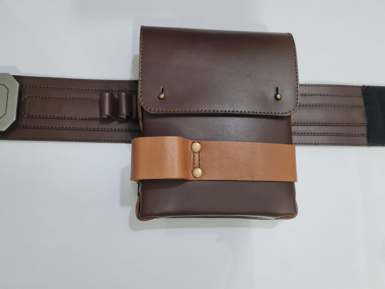 Puede incluir: Un cintur&oacute;n de cuero marr&oacute;n con una bolsa a juego y una correa marr&oacute;n m&aacute;s clara. La bolsa tiene un cierre de solapa con remaches de lat&oacute;n. El cintur&oacute;n tambi&eacute;n presenta una hebilla plateada y peque&ntilde;os bucles.