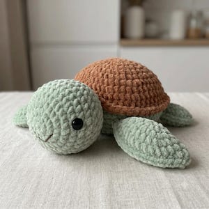 Op de afbeelding: Een pluche schildpadspeeltje gemaakt van zacht, grof garen. De schildpad heeft een afgeronde, getextureerde lichtgroene kop en flippers, gecombineerd met een opvallend, koepelvormig bruin schild. Het heeft één zichtbaar zwart plastic oog.